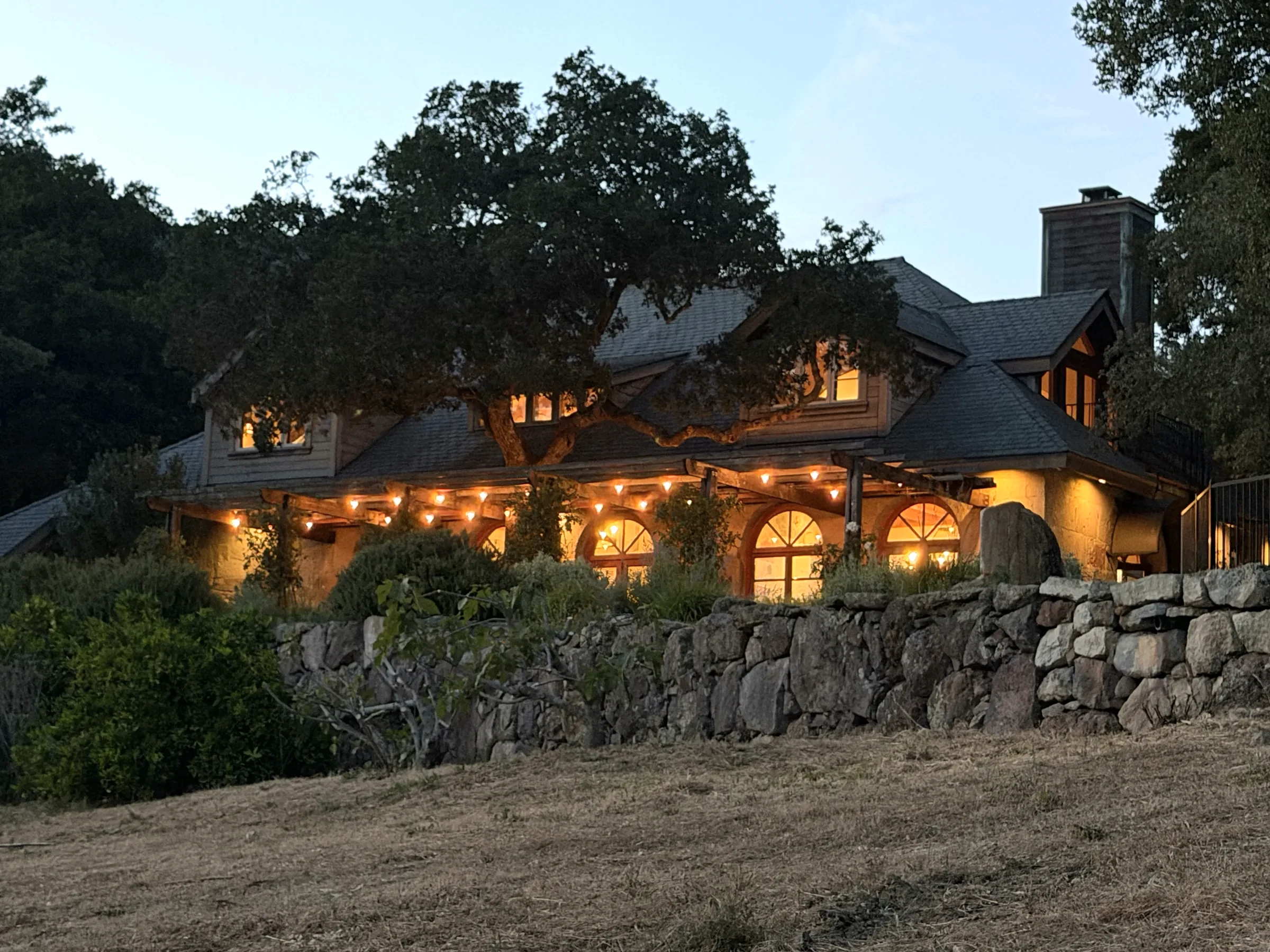 The Stone House at dusk, string lights under the pergola, the stone wall below